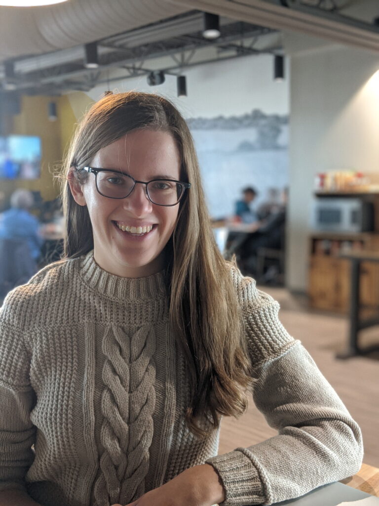 Emily is sitting in a coffee shop wearing a hand-knit sweater. She is smiling widely. Her glasses are slightly crooked.